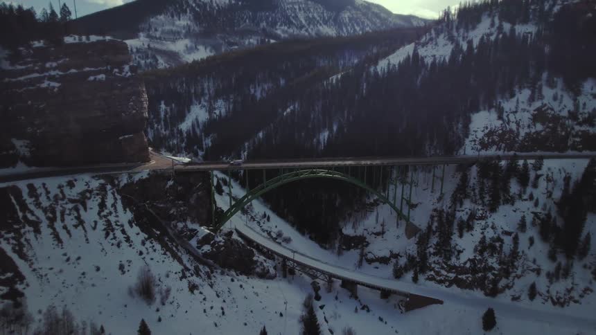 An aerial view of a bridge with a street above where a car passes and there are hills full of trees and snow on the ground