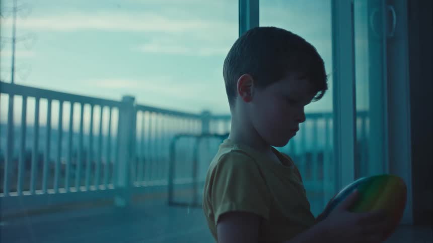 A child playing with a ball while leaning against the glass indoor at sunset