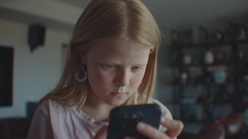 A little blonde girl is looking at the cell phone that is in her hand and then she closes her eyes and opens them after a while