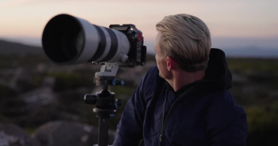 A man outdoors with a camera on a tripod capturing the landscape at dusk