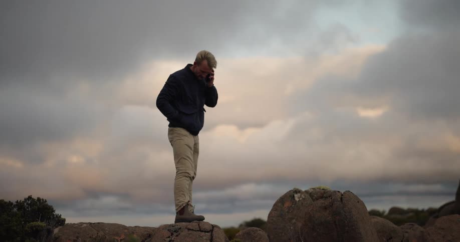 A man is standing on rocks along the coast looking around as he answers his phone
