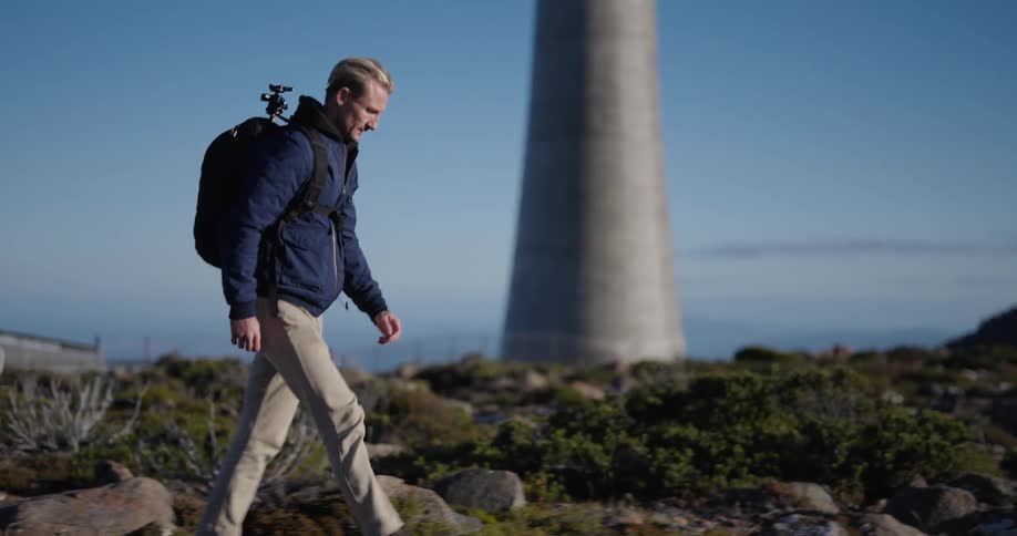 A person with a backpack and camera equipment is walking near a tall structure amidst rocky terrain under a clear sky