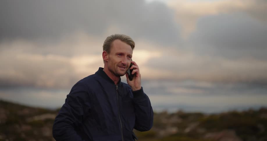 A blond man is talking on the phone while his hair is moving in the wind