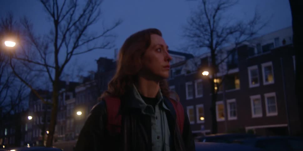 A fair, red-headed woman glances to the side and ahead at her surroundings as she walks down a lit city sidewalk at dusk.