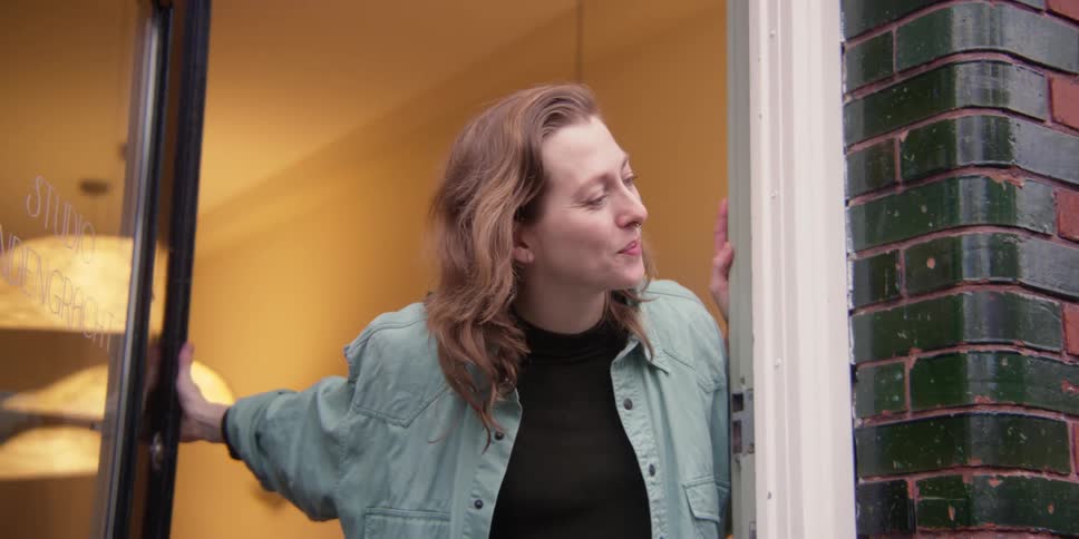 A fair, light-haired woman waves goodbye as she lets a young adult male with curly hair and leather coat out the front door of her windowed studio.