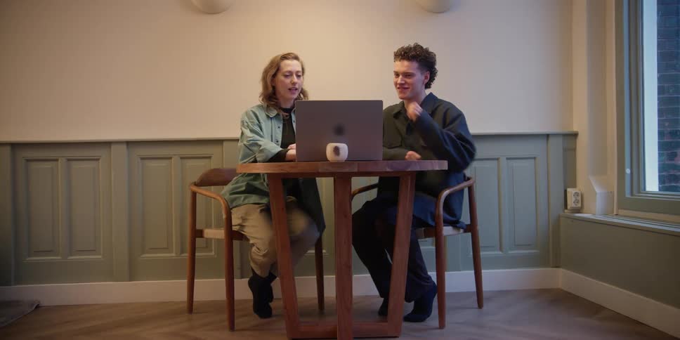 An adult red-headed  woman and young adult male with curly dark curly hair pull the chairs out and sit at a table, as the woman puts her laptop down, and then they both lean in and interact over what they are viewing on the screen.