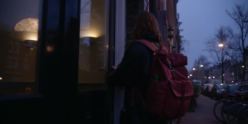 A fair red-headed woman walks on a city sidewalk before sunrise with rows of parked bikes and streetlights, then turns toward the door of a windowed, brick business to unlock it and let herself in.