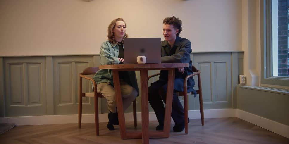 An adult woman and young adult man sit at a table together with a laptop in front of them and expressively discuss what they are viewing.