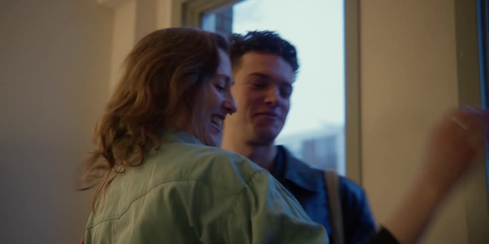A red-headed middle-aged woman hugs a young curly-haired adult male as he enters her studio with windows to the city outside.
