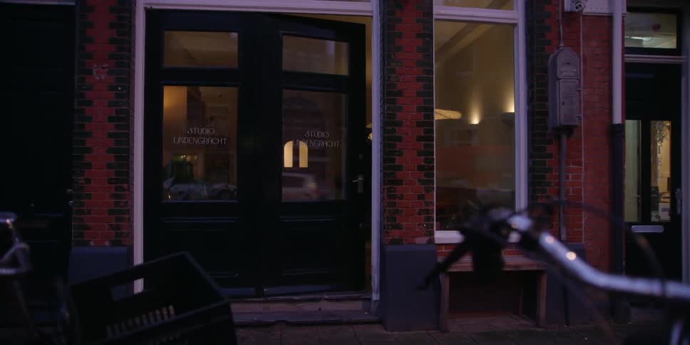 POV behind bicycle handlebars of a woman walking on the sidewalk with a backpack on as she turns to a windowed studio, unlocks the door, goes inside, and turns on the lights.