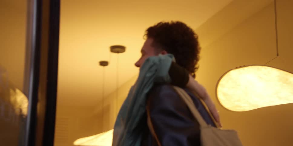 A young curly-haired man stands outside a windowed studio door and is let in and hugged by an adult woman with red hair.