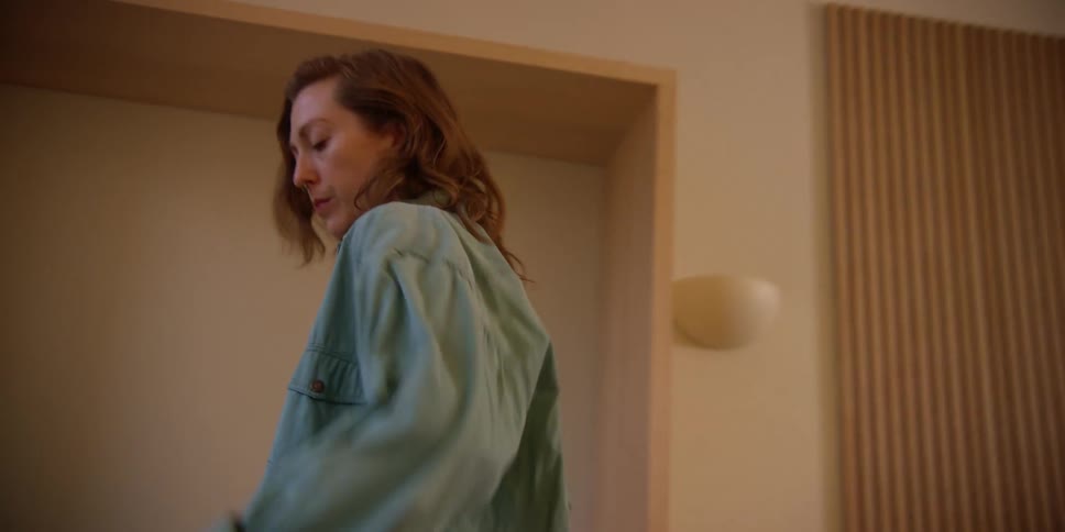 A white, red-headed woman shuffles her feet and moves her arms in smooth motions as she dances in her living room with a far off expression.