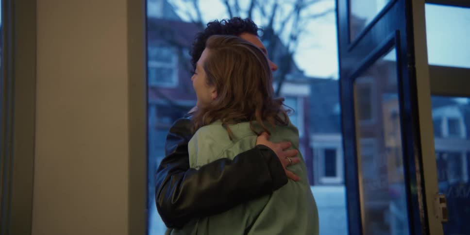 An adult woman with red hair opens the windowed store-front door to greet and hug a young adult male with dark curly hair who comes inside.