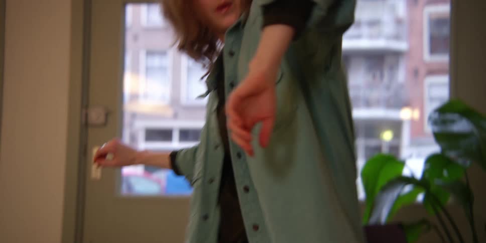 A red-headed female dancer practices arm and leg movements for a contemporary dance in front of the windows to the city outside.
