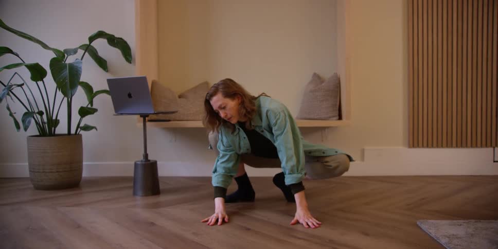A red-headed middle-aged woman is doing stretches in her living room with a computer on a stand behind her.