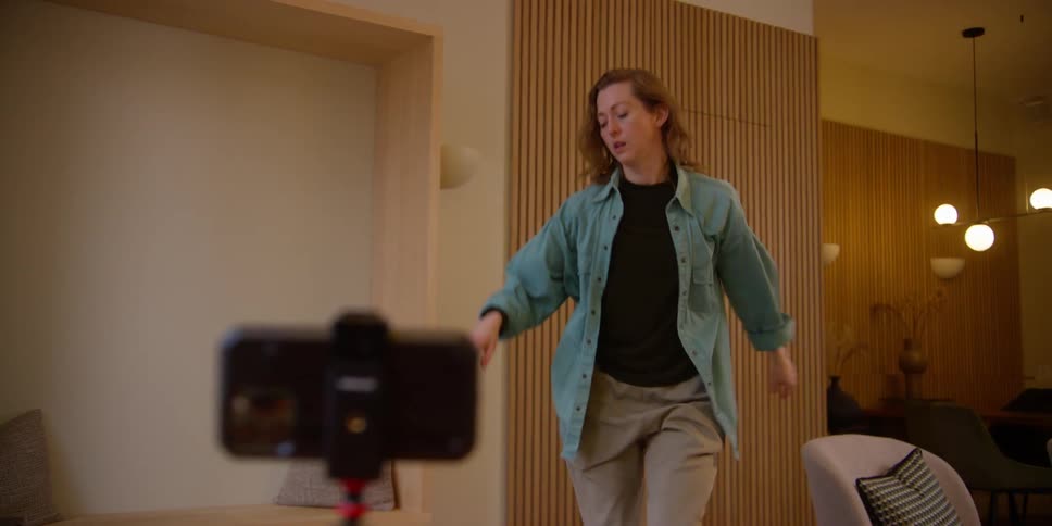 A fair, red-headed female dancer records herself practicing choreography with a smartphone on a tripod and then turns off the camera