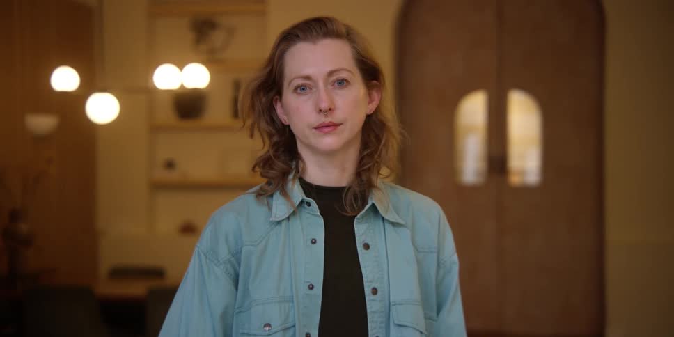 A red-headed woman with a septum ring standing in a mid-century style room is looking away but then turns to the camera with a blank expression