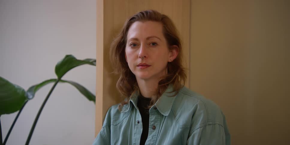 Portrait of a fair-skinned woman with auburn hair and a nasal piercing leaning against a wooden cabinet indoors.