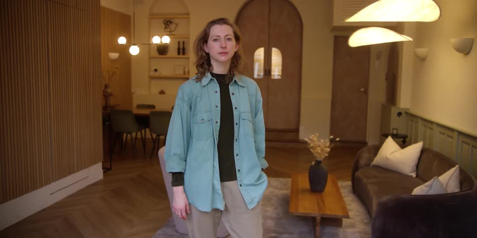 A tall white woman with ginger hair and septum nose piercing is slightly smiling and standing with hand in pocket in a retro living space.