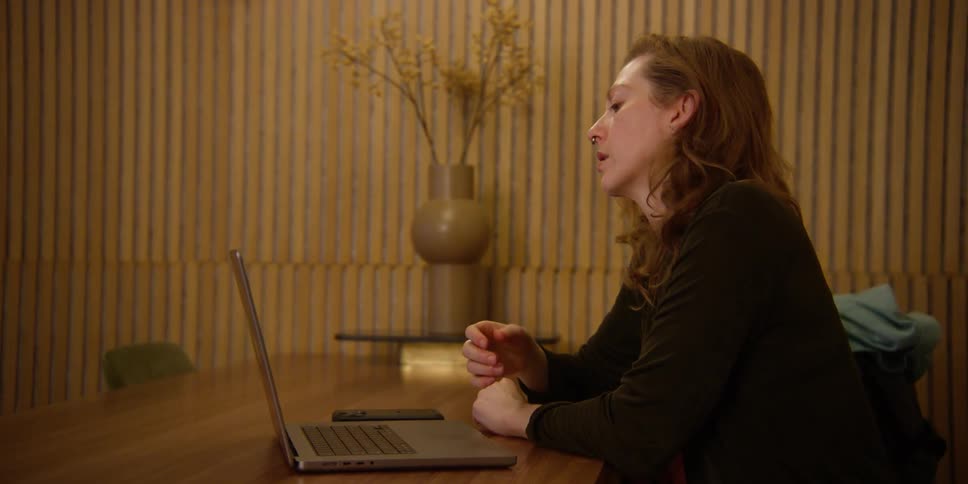 A woman with fair complexion and red hair studies dance choreography on her laptop and practices a movement with her hand.