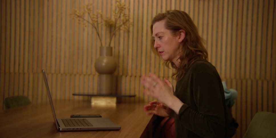 Woman sitting at an office desk watching a dance routine on her laptop and emulating some of the moves with her body and hands.