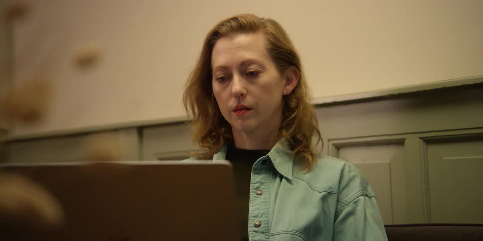 View from behind a plant of a woman with fair skin, auburn hair, and an open jean button-down shirt working on her laptop.