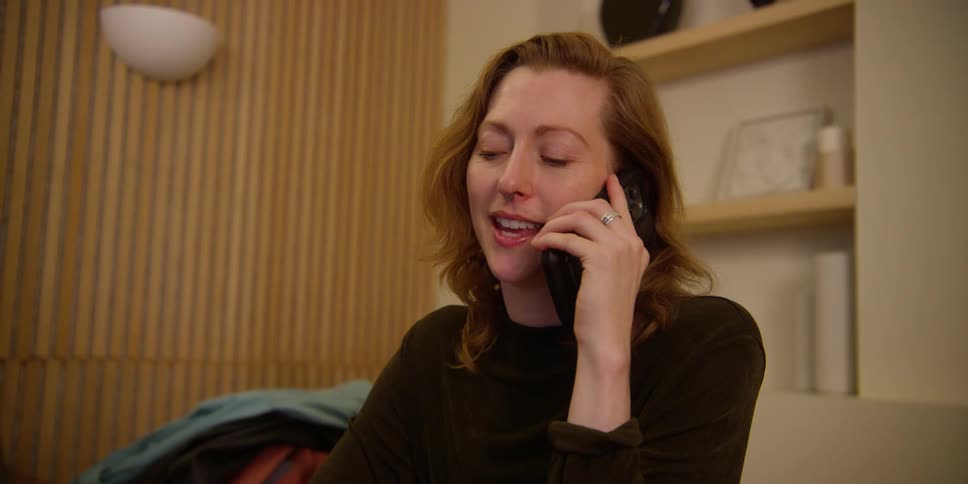 A woman with light-skin and red hair gestures and emotes with facial expressions as she talks on her cell phone in her office.