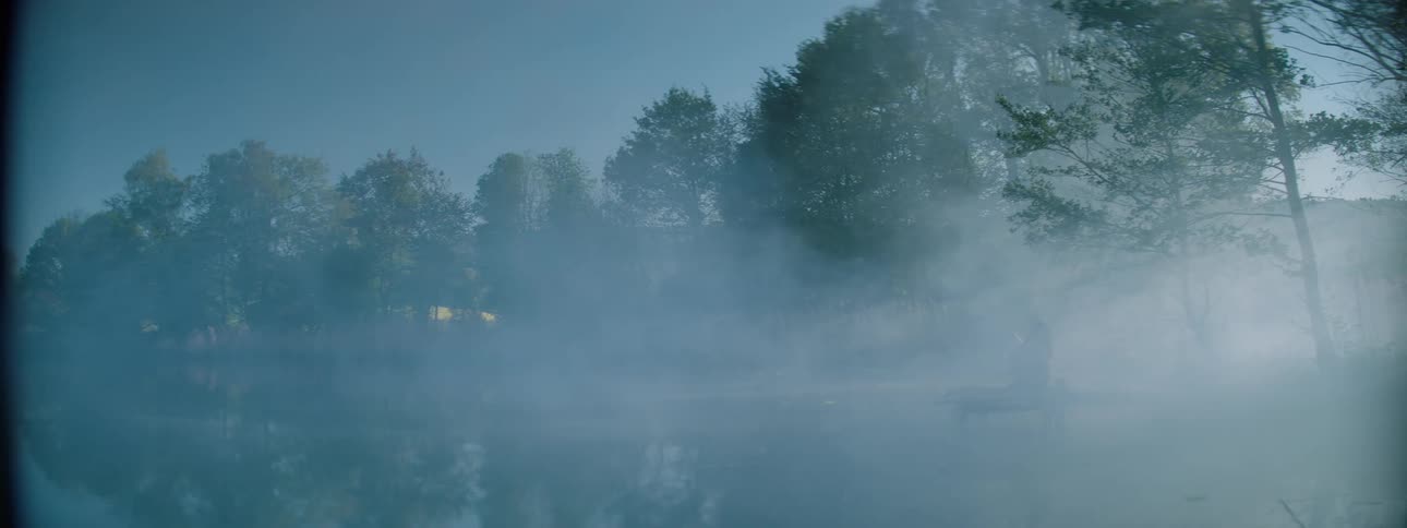 A person sitting by the river bank as fog cover the whole atmosphere with trees surrounding the lake