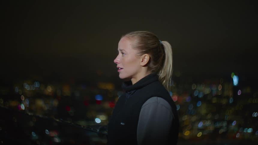 A blonde woman is looking to the left while breathing tiredly and behind her there are many lights of a city in a night