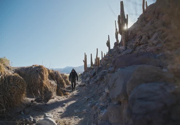 A person in warm clothes is walking along a path surrounded by rocks and cactus in a desert on a sunny day