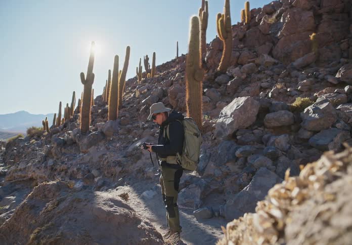 A man walking along a desert path stops to look through his binoculars then moves on