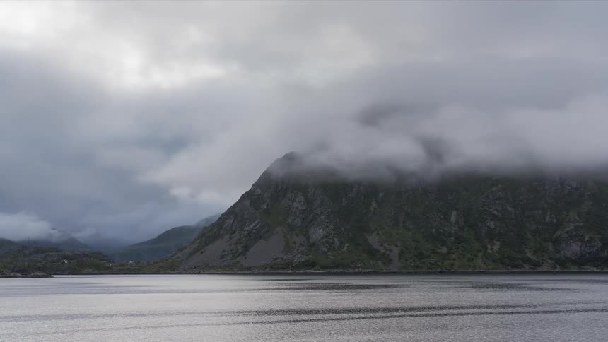 A mountain facing the sea that is moving to the right and there is a large cloud passing over the mountain peak