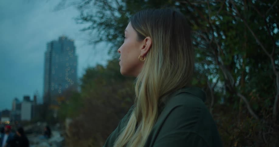 A woman in a city park breathes deeply with her eyes closed then opens her eyes and looks around