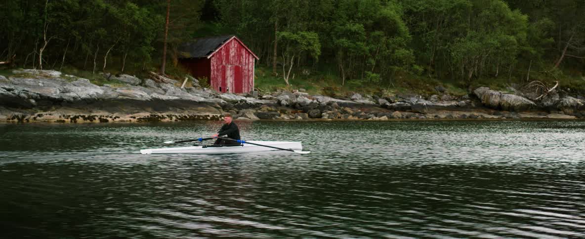 Man rowing a kayak on a calm lake past an iconic red Norwegian fishing hut