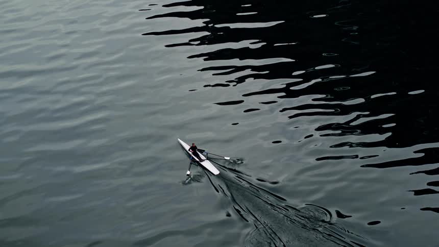 A person rows a stark boat across a high contrast glassy surface on a lake with a moody feel