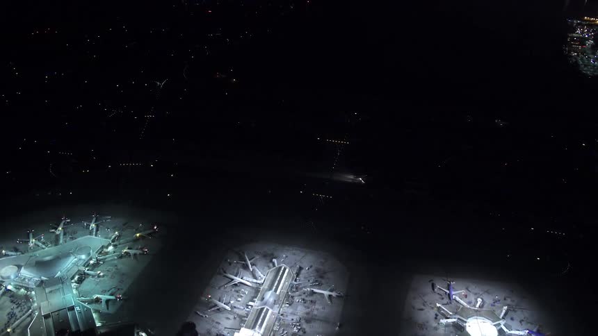 A bird's-eye-view of an airport showing several terminals with parked planes and a plane moving at night