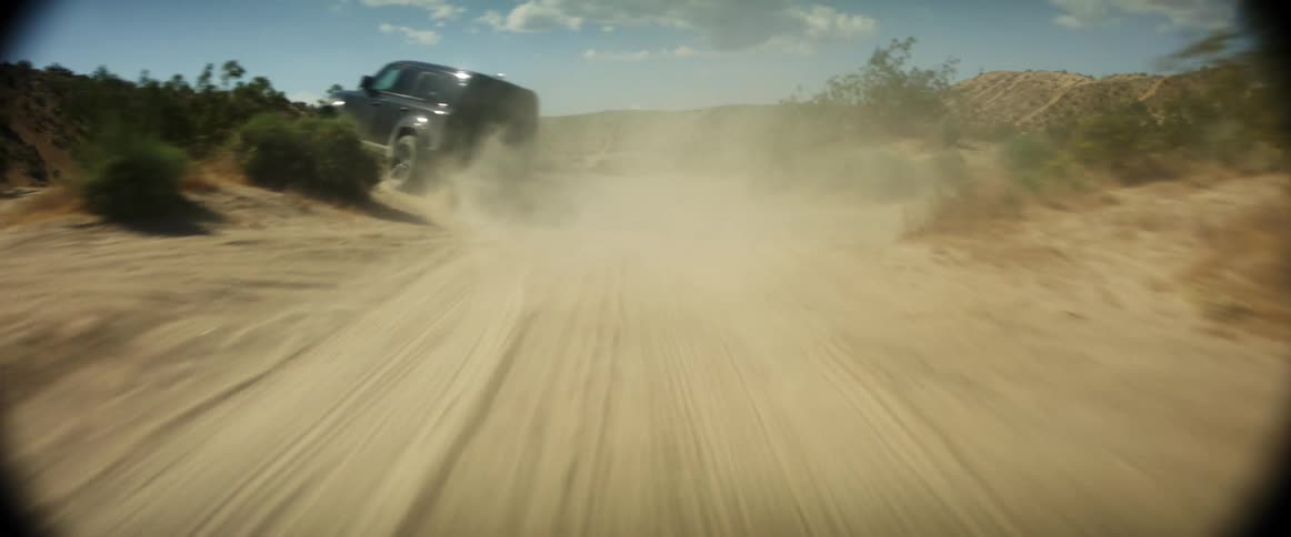 A car travels quickly down a dirt road in a desert environment kicking up a cloud of dust behind it