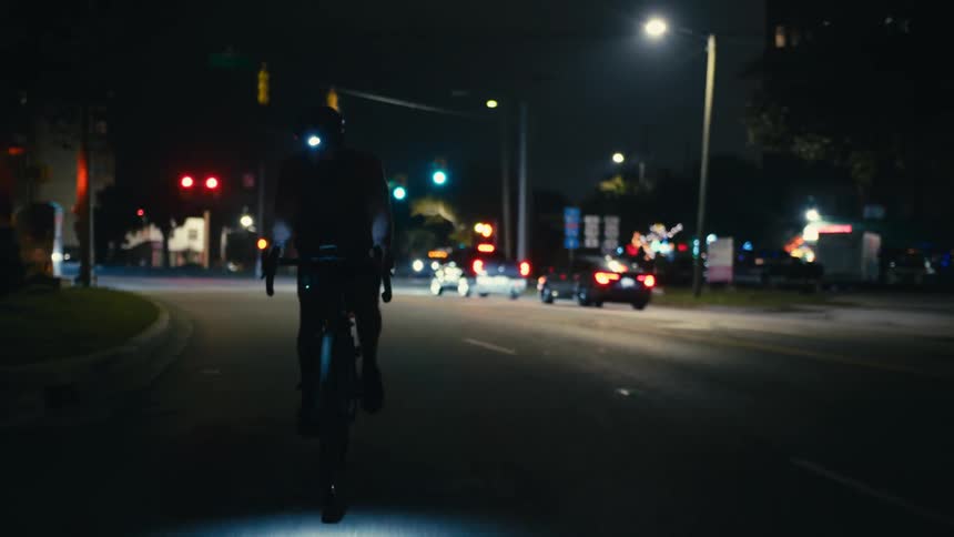 A man in a helmet with a flashlight pedaling his bicycle through the city streets