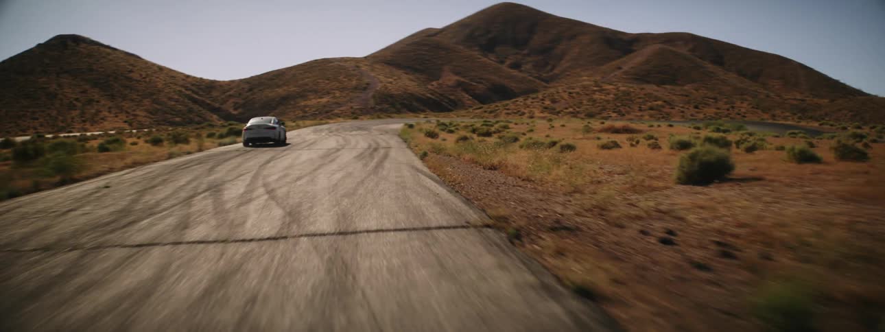 A white car is speeding on a desert road with mountains visible in the distance