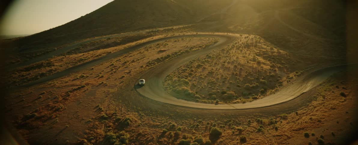 White car drives down a steep curved road in front of a mountain