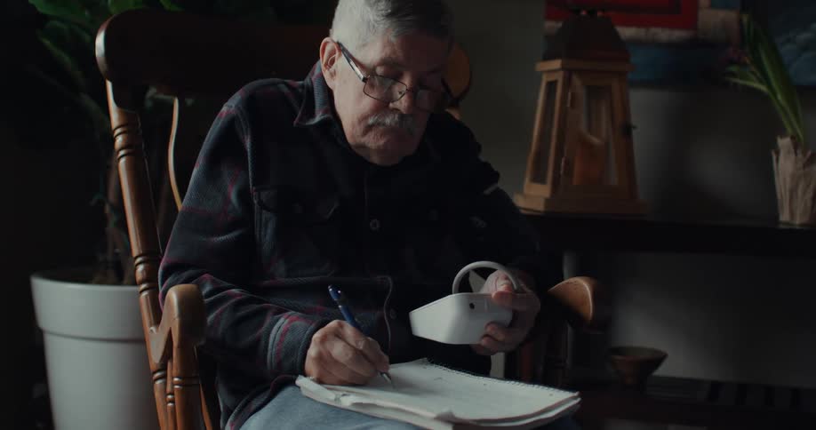 An elderly man is seated writing in a notebook with a looking at a small electronic device