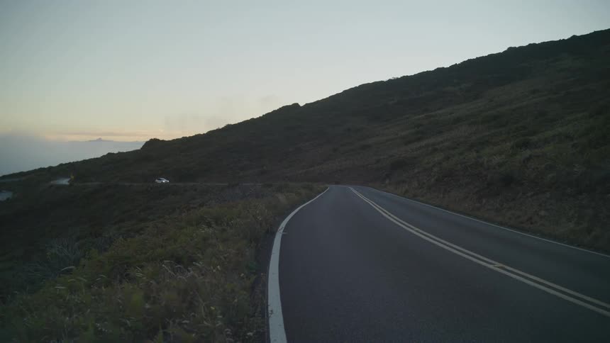 A car travels on a winding road amidst hilly terrain during sunset
