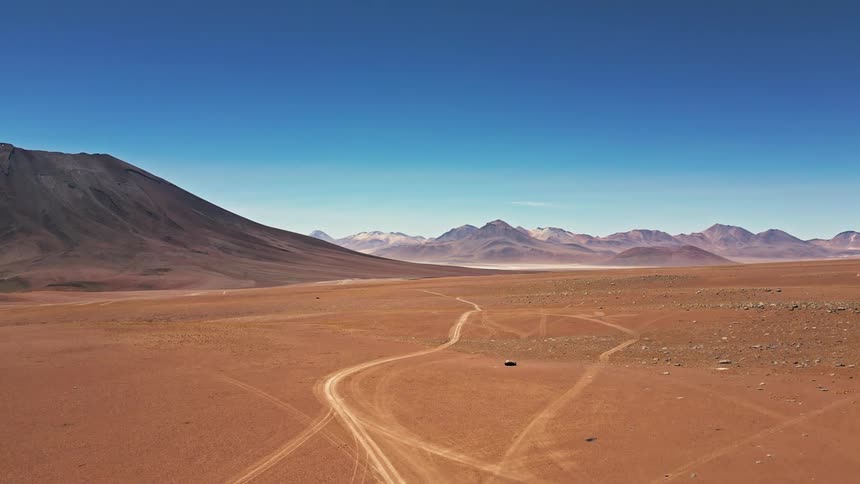 A vast barren desert landscape with tire tracks crisscrossing the sandy soil stretching toward distant mountains under a clear blue sky