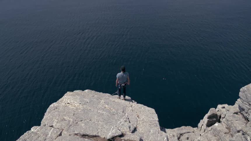 Man stands up after reaching the top of a rocky mountain lining the ocean