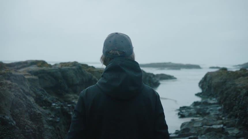 Man in cap and jacket walking towards the sea with rocks around