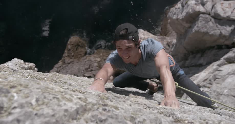 A man is climbing a mountain using a rope and he blows on his hand and dust comes out and down to a sea coast with the rock he is climbing