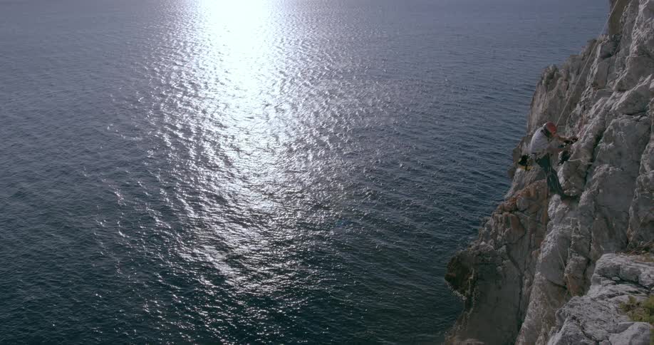 A rock climber going up a cliff over the ocean throwing down his security rope