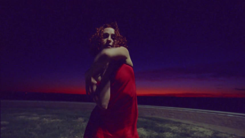 Woman with red curly hair dancing in an open environment wearing a red dress on a night with a red sky