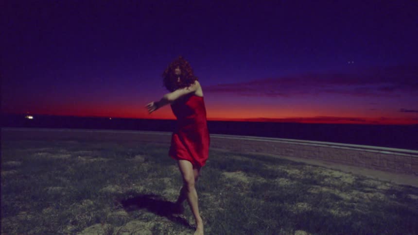 A woman in a red dress dances strangely in a grassy field with an orange sunset behind her