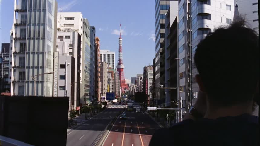 A persons back is taking a photo of a city street as vehicles pass by and in the background there is a red and white tower